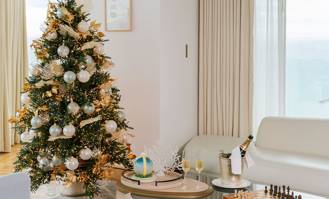 A luxuriously adorned Christmas tree with golden accents and soft lights stands in a bright, minimalist living room with champagne and elegant decor on a nearby table.