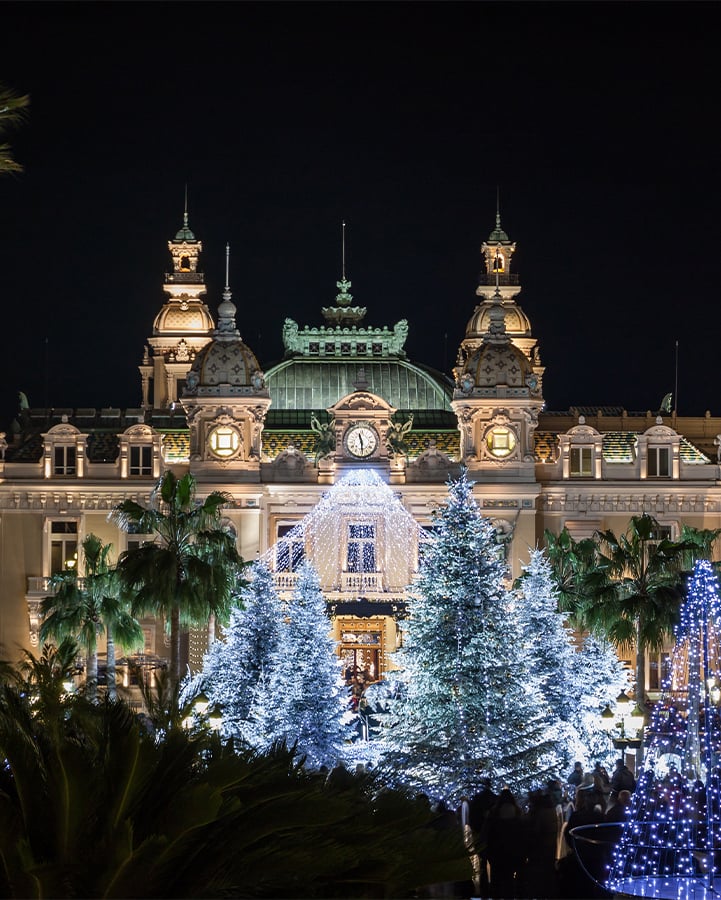 A majestic building adorned with elegant lighting and surrounded by sparkling, snow-covered Christmas trees creates a magical festive atmosphere at night.