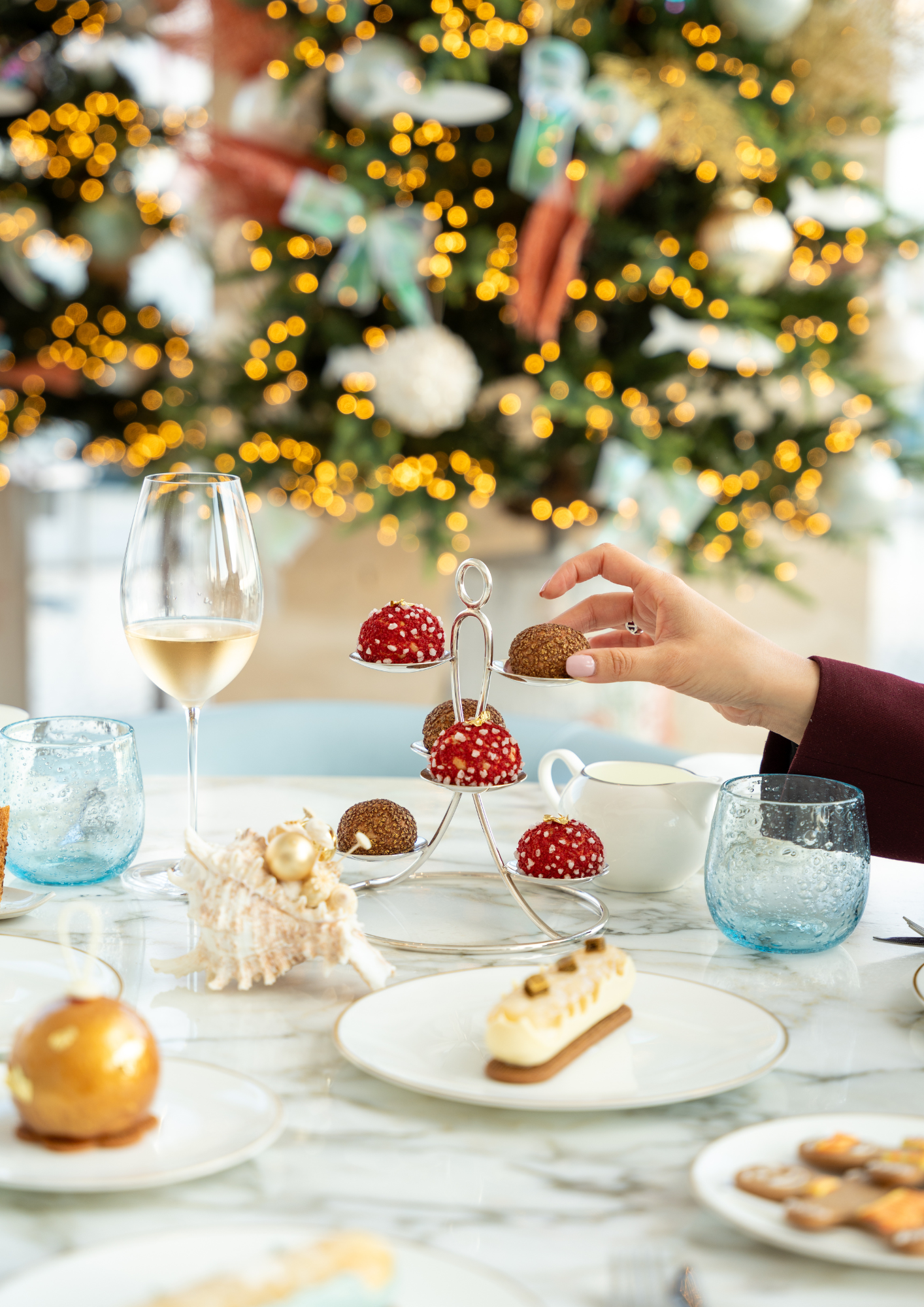 Un tea time au restaurant Riviera sur le thème de Noël, avec une main qui attrape une mignardise rouge aux couleurs de Noël et une coupe de champagne posée sur la table. An afternoon tea at the Riviera restaurant with a Christmas theme, featuring a hand reaching for a red petit four in festive colours and a glass of champagne on the table.