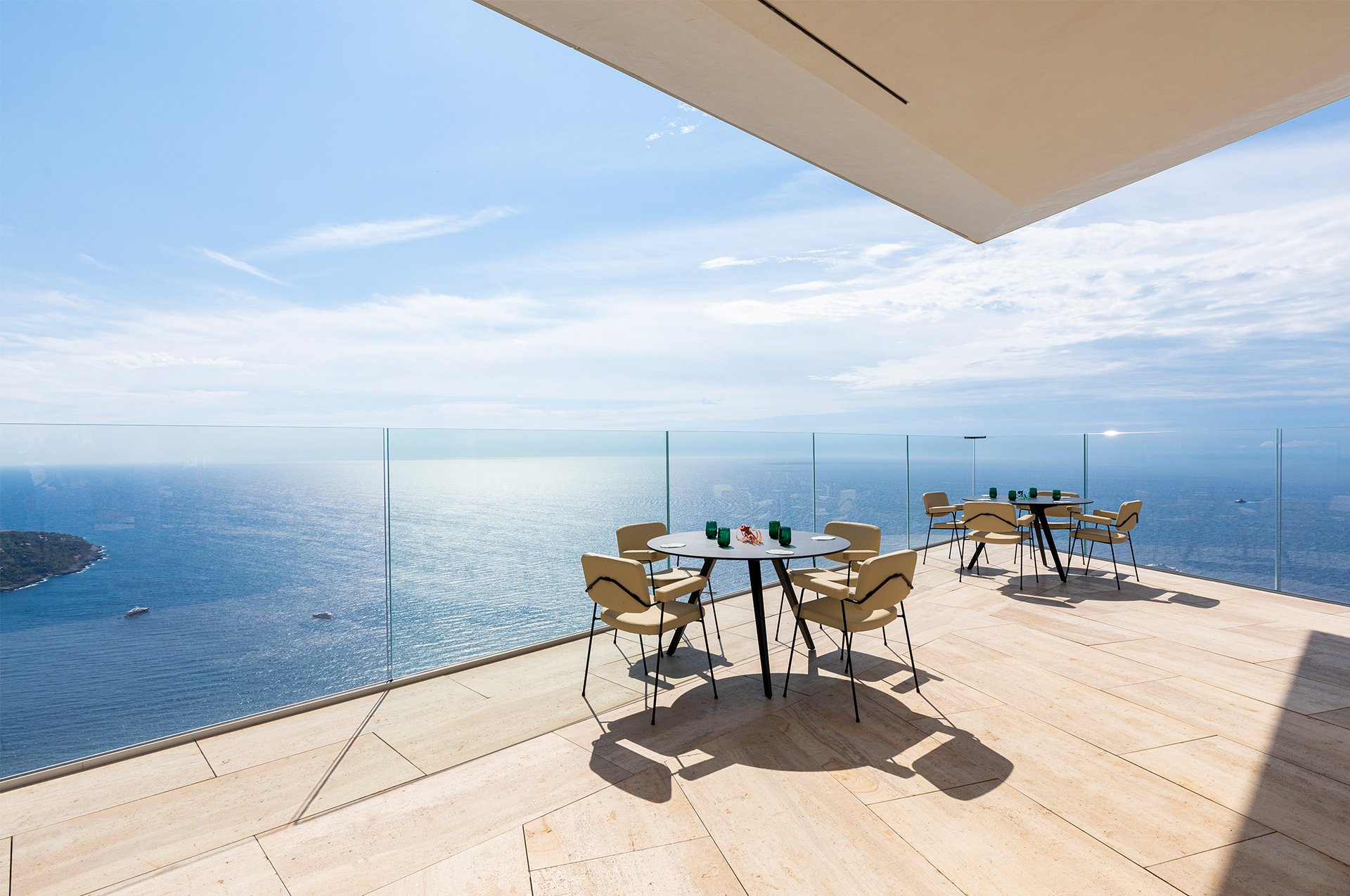 A panoramic terrace with modern dining tables and chairs, offering breathtaking views of the shimmering sea and expansive horizon.