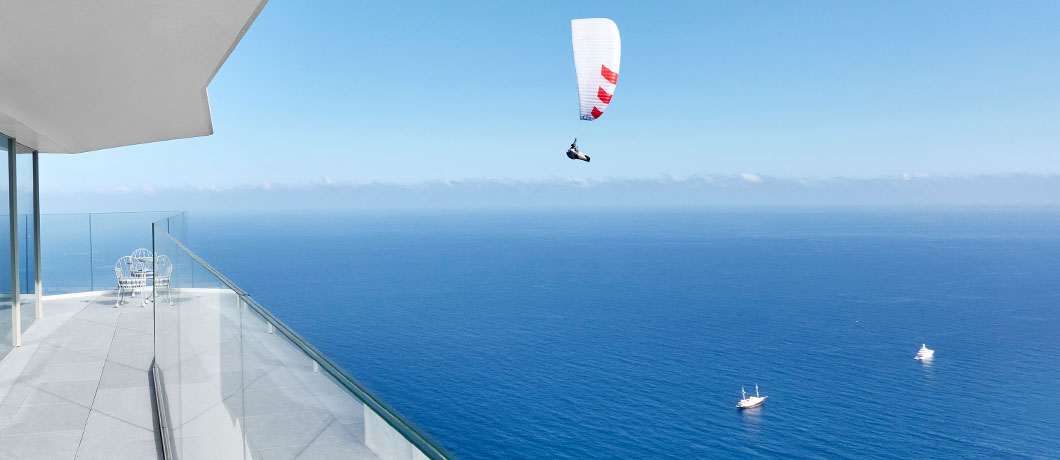 View of person paragliding over the sea, with a boat in the distance. A balcony at The Maybourne Riviera is also in view.