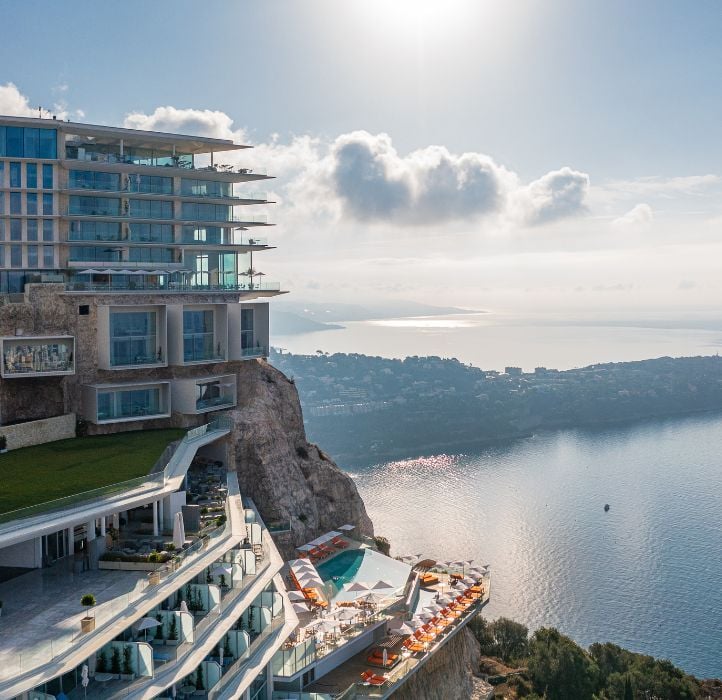 The luxurious, The Maybourne Riviera building is perched on a cliffside, offering stunning views of the ocean and surrounding landscape, with a pool area on a lower terrace. The sky is clear, with sunlight reflecting off the water below.