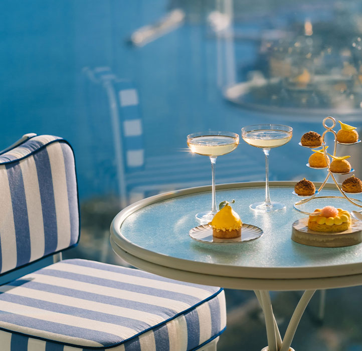 A table set with elegant desserts and cocktails overlooks a scenic, blue view. The setting includes striped blue and white cushions, creating a relaxed, luxurious atmosphere.