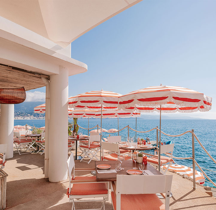 An elegant seaside restaurant with orange and white striped umbrellas and cushioned chairs overlooking a sparkling blue ocean. The setting features a sunny, clear day with a beautiful view of the coastline.