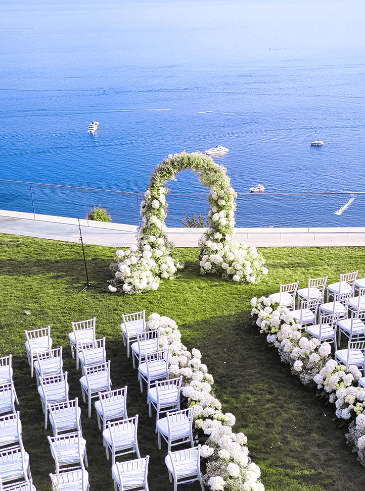 A breathtaking oceanfront wedding ceremony setup featuring a floral arch of white blooms, neatly arranged white chairs, and panoramic views of the deep blue sea with yachts in the distance.