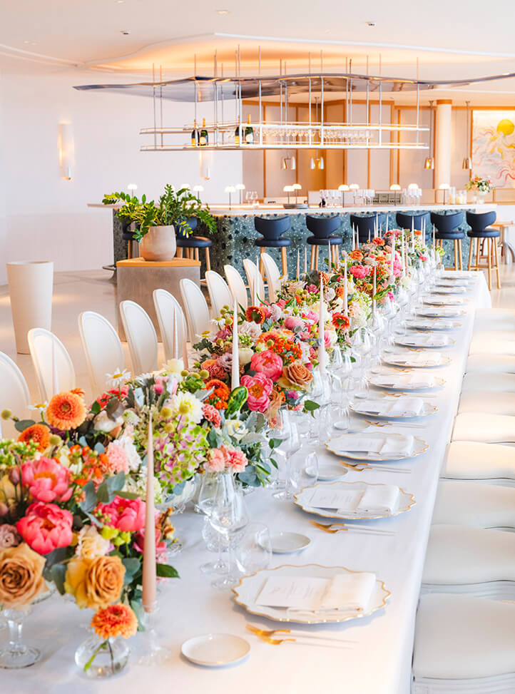 A beautifully set private dining table adorned with vibrant floral arrangements, elegant gold-rimmed plates, and tall candles, set against a contemporary bar in a refined venue.