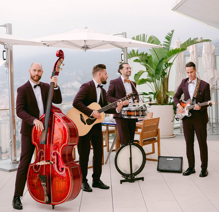 A four-piece band, dressed in matching maroon suits with bow ties, performs on an outdoor terrace. The musicians play various instruments, including a double bass, acoustic guitar, snare drum, and electric guitar, with a scenic view of mountains and the city in the background.