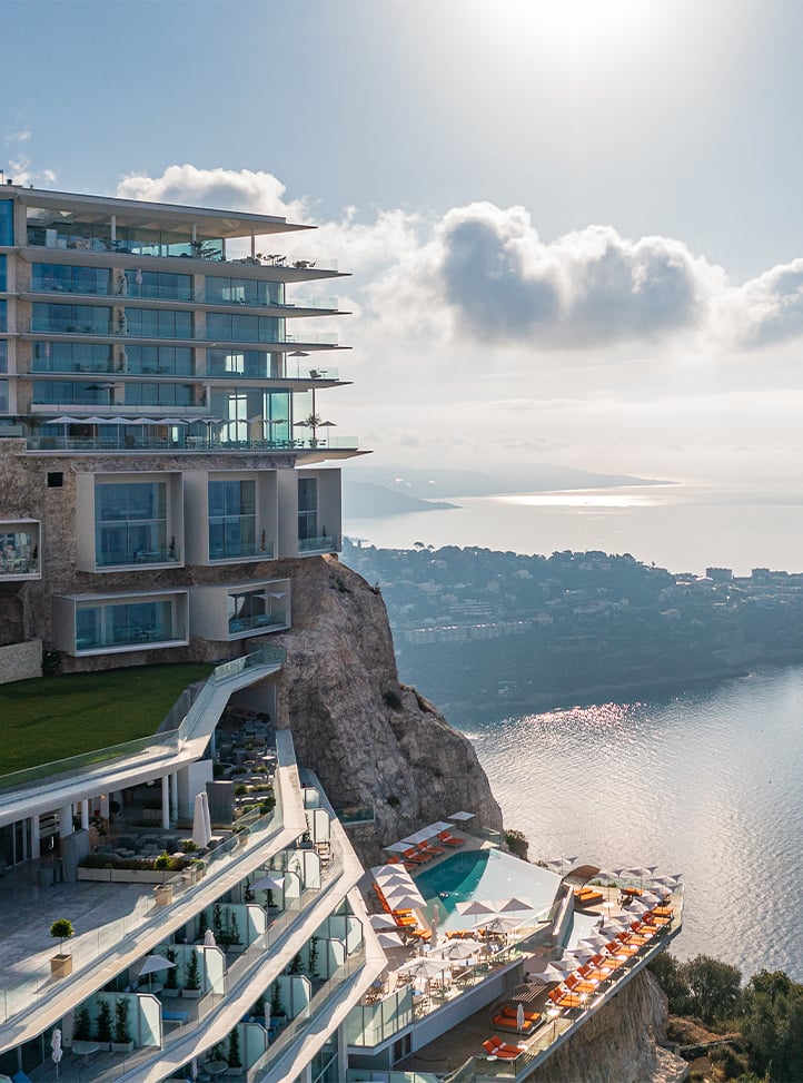 An aerial view of The Maybourne Riviera hotel, perched dramatically on a cliff overlooking the Mediterranean Sea. The modern, multi-level glass structure features terraces, pools, and stunning panoramic views of the coastline and surrounding areas.