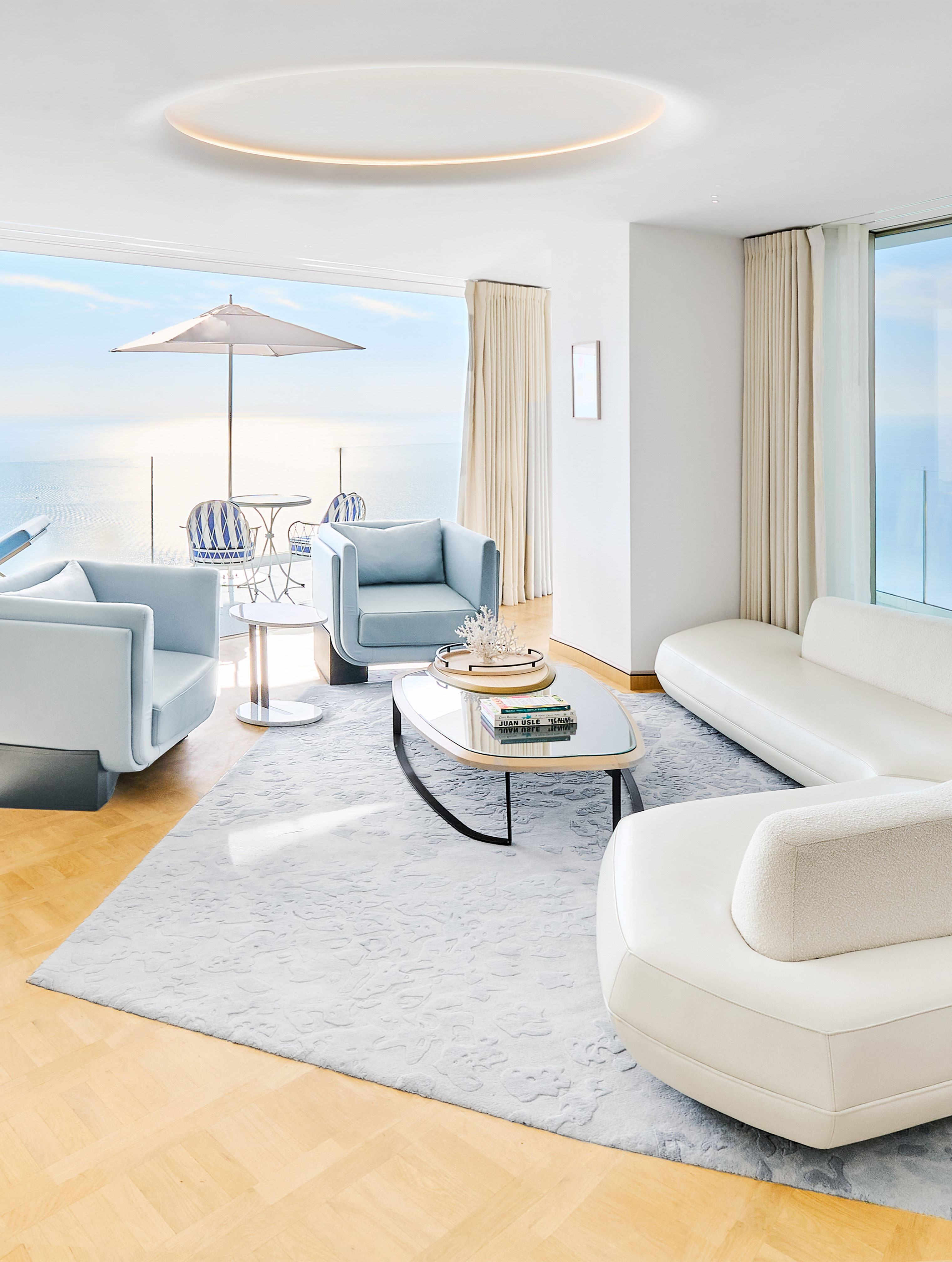 This image depicts a bright, modern suite with light blue and white furnishings, featuring a seating area with armchairs and a sofa, a coffee table, and an outdoor balcony with a stunning view of the sea. The decor is elegant and minimalistic, with large windows that allow for ample natural light.