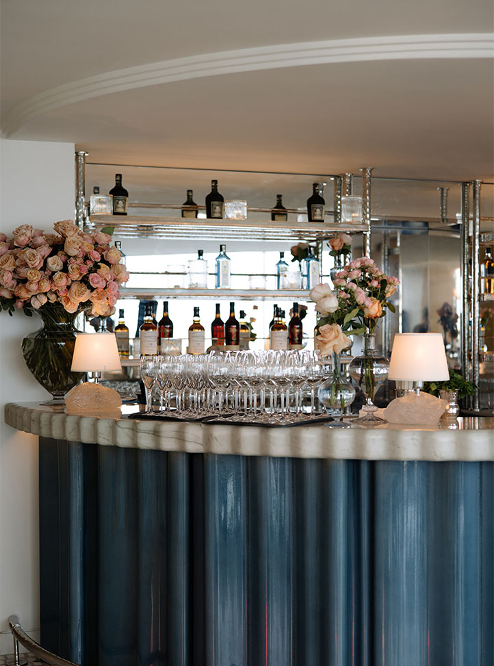 A stylish bar setup with a sleek marble counter, lined with glassware and decorated with elegant floral arrangements. Shelves behind display a selection of premium spirits, creating a sophisticated ambiance for guests.