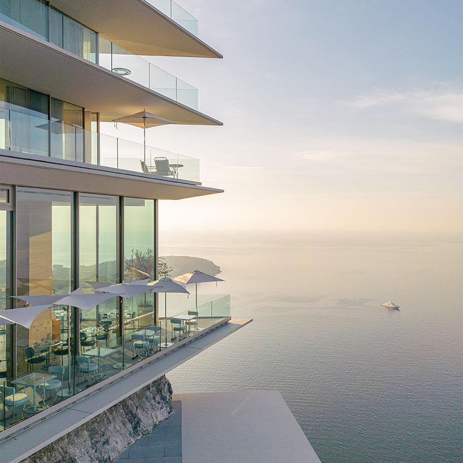 The image shows a modern, minimalist building with glass balconies overlooking a serene body of water, possibly the ocean. The structure appears to be suspended on a cliff edge, providing an expansive and breathtaking view of the horizon.