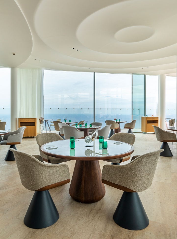 A sunlit dining area with contemporary, rounded chairs in soft hues, surrounded by glass walls showcasing sweeping seaside vistas, exuding modern refinement.