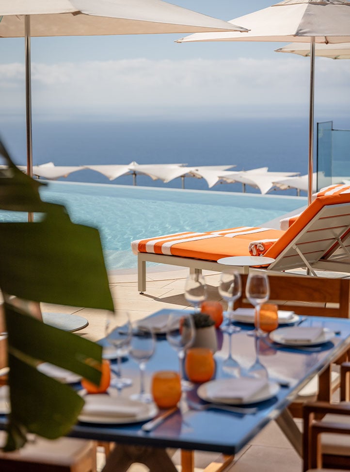 A chic poolside dining setup with vibrant orange lounge chairs, an inviting infinity pool, and a backdrop of the endless blue horizon, perfect for leisurely luxury.