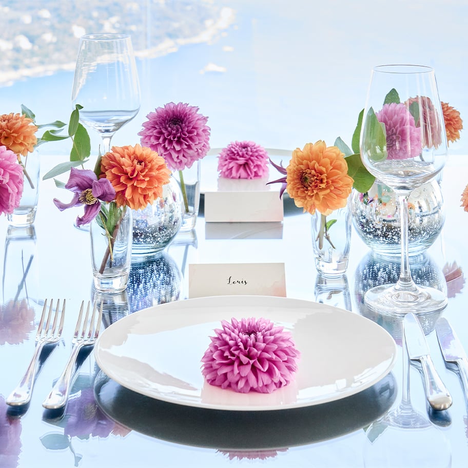 A sophisticated dining table set with delicate floral arrangements in soft pinks and oranges, reflecting against a polished surface with a stunning coastal backdrop.