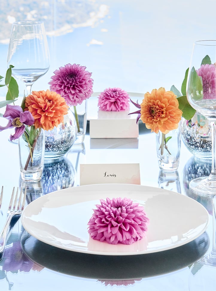 A sophisticated dining table set with delicate floral arrangements in soft pinks and oranges, reflecting against a polished surface with a stunning coastal backdrop.