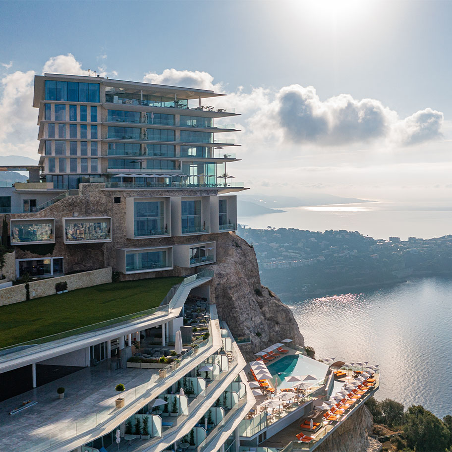 An aerial view of The Maybourne Riviera hotel, perched dramatically on a cliff overlooking the Mediterranean Sea. The modern, multi-level glass structure features terraces, pools, and stunning panoramic views of the coastline and surrounding areas.