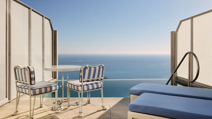 A small terrace with blue-striped chairs and a table beside an infinity pool, offering a clear view of the ocean horizon.