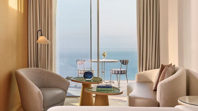 A cozy sitting area with modern furniture inside a suite, facing a balcony with a table and chairs overlooking the sea.