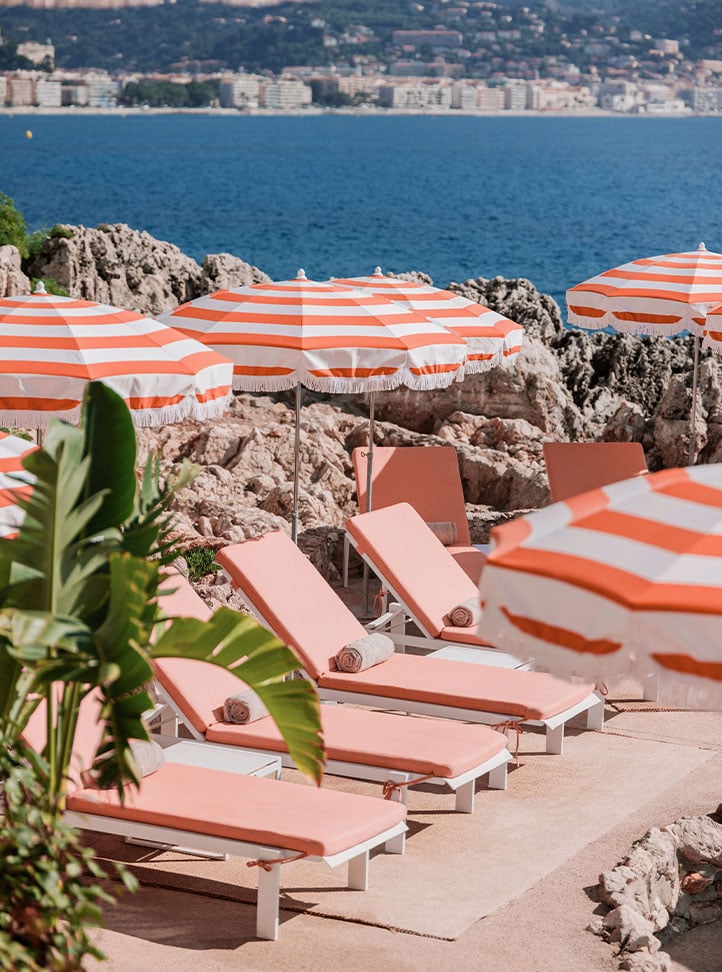 Peach coloured sunbeds, with a towel on each, and striped parsols. In the background is the sea, greenery and buildings.