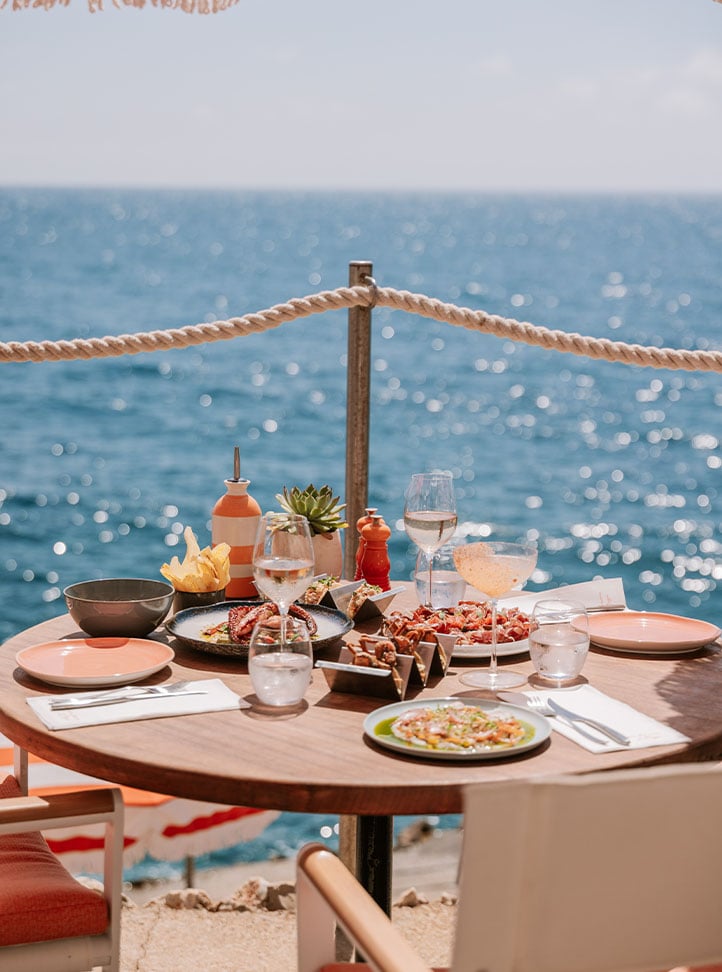 A table at Maybourne La Plage, facing the sea. On the table is a selection of food, 2 glasses of rose and a cocktail.