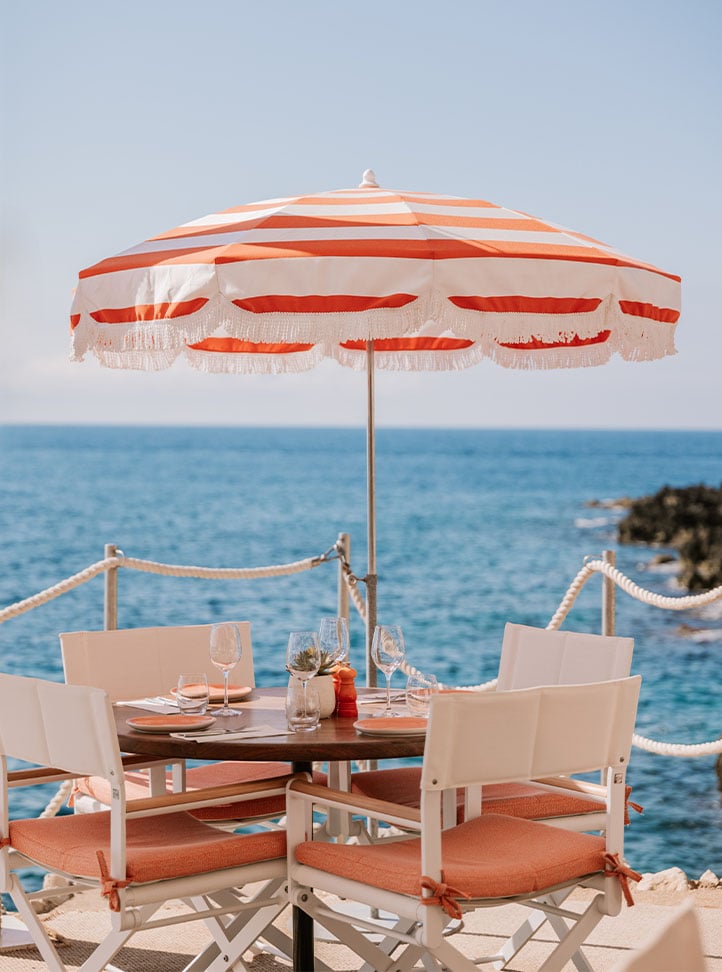 Table at Maybourne La Plage, by the sea. The table is laid up with cutlery, plate and wine glasses. A striped parasol creates shade for the table.
