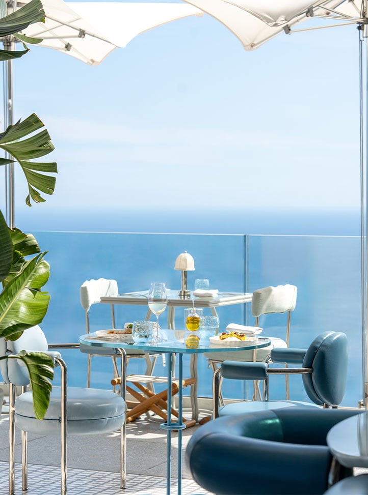 tables on terrace laid up with wine glasses, cutlery, plates and napkins. View over sea and blue sky.