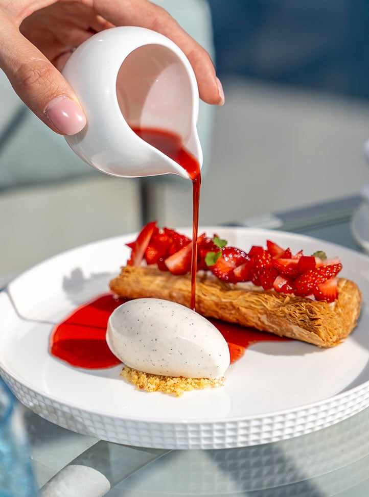 hand pouring pink sauce onto a strawberry orange blossom mille-feuille on a white plate.