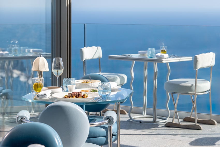 Terrace table with steak and wine glasses, blue sky, another table in the background and a sea view.