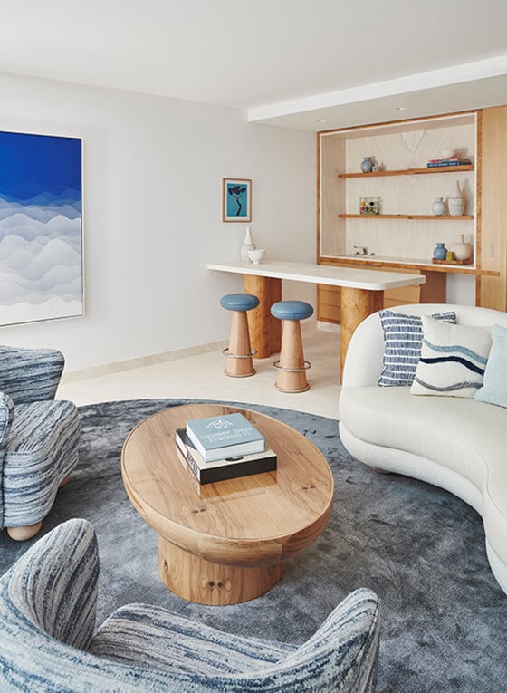 The Maybourne Suite living area, with 2 arm chairs, coffee table with books, a sofa, a table with two stool tucked behind it and at the back of the room is a shelf with ornaments scattered across the layers.