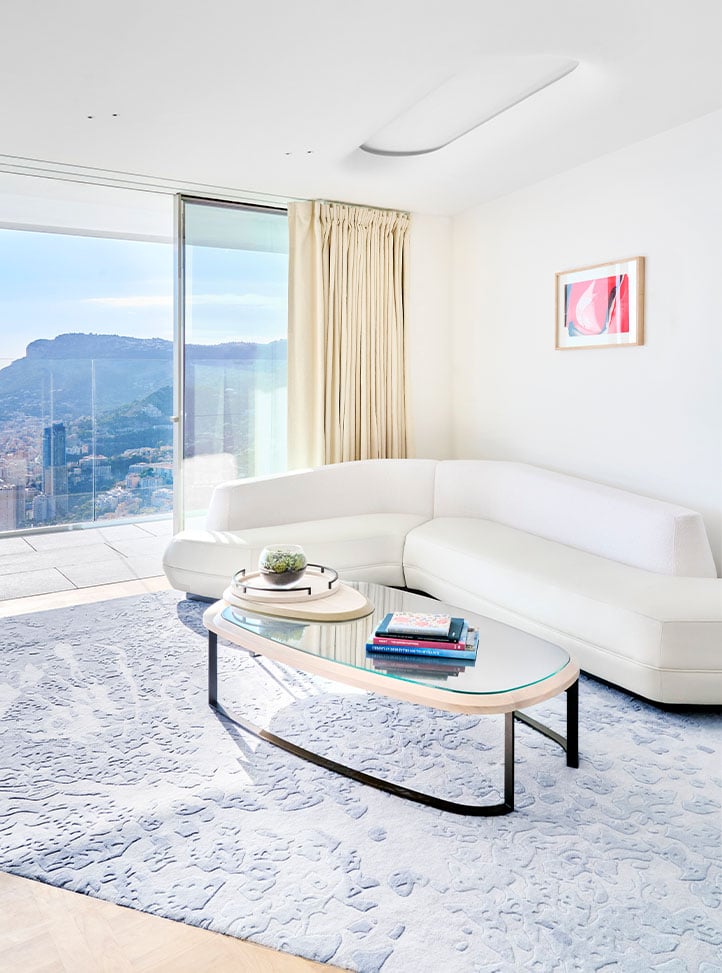 A modern living room in a suite with a curved white sofa, a glass coffee table, and a large window offering a scenic view of a mountainous landscape.