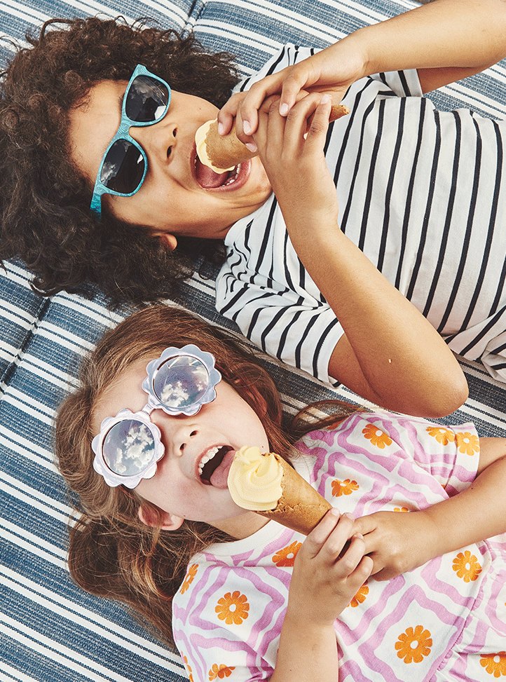 2 children, girl on the left and boy on the right, lying on a blue & white striped sunlounger, laughing with an ice-cream each in hand.