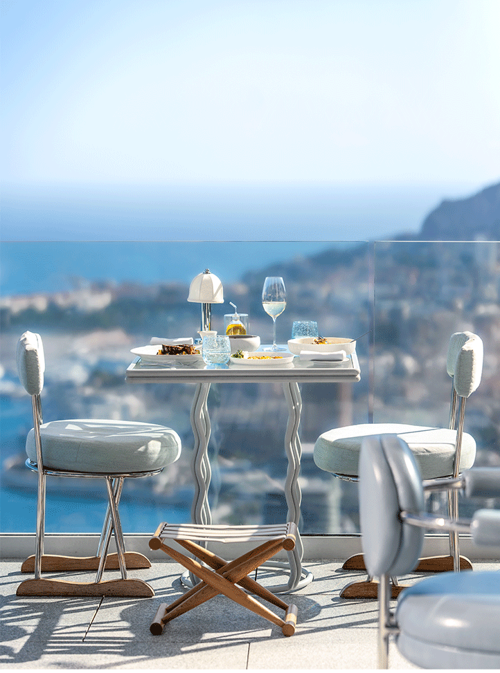 A beautifully set outdoor dining table with modern chairs overlooks a stunning ocean view. The table is elegantly arranged with plates of food, a glass of white wine, and a small lamp, creating a serene and luxurious atmosphere.