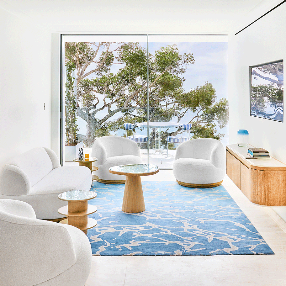 A bright, modern suite with white, curved furniture and a blue patterned rug. The space features large sliding glass doors that open to a balcony with outdoor seating, overlooking lush greenery and a serene view.