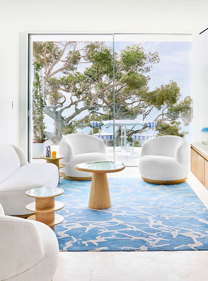 A bright, modern suite with white, curved furniture and a blue patterned rug. The space features large sliding glass doors that open to a balcony with outdoor seating, overlooking lush greenery and a serene view.