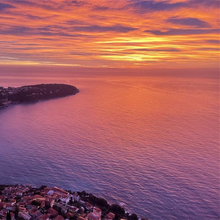 A breathtaking aerial view of the French Riviera at sunset, with the sky painted in vibrant hues of orange, pink, and purple, reflecting over the calm Mediterranean waters, as the coastline curves elegantly around a secluded bay.