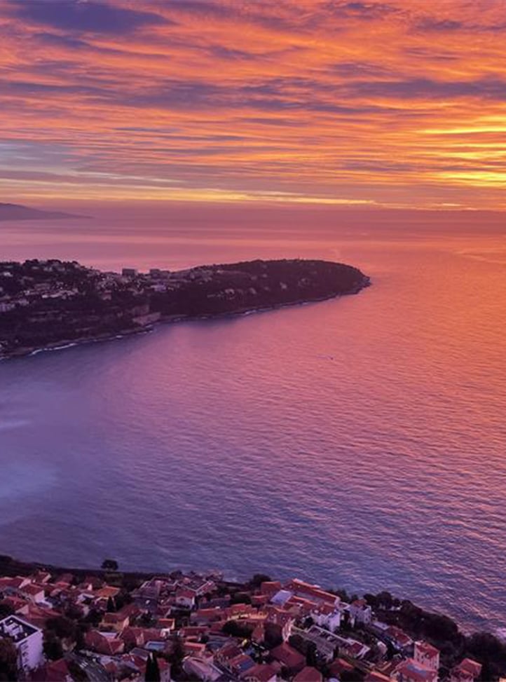 A breathtaking aerial view of the French Riviera at sunset, with the sky painted in vibrant hues of orange, pink, and purple, reflecting over the calm Mediterranean waters, as the coastline curves elegantly around a secluded bay.