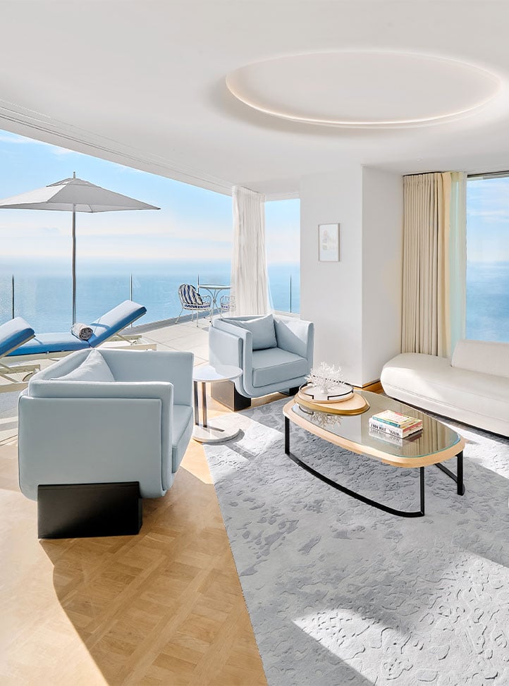 Claridge's suite with coffee table, coffee table books, a white sofa and two pale blue arm chairs. In the background is a balcony with sunbeds and a view of the sea.